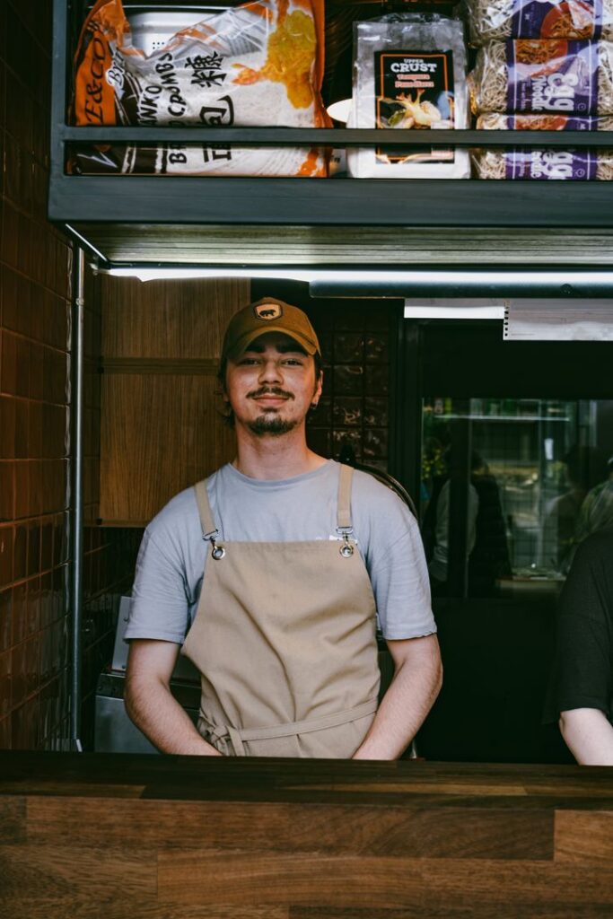 A smiling barista stands confidently in an urban cafe wearing a cap and apron.