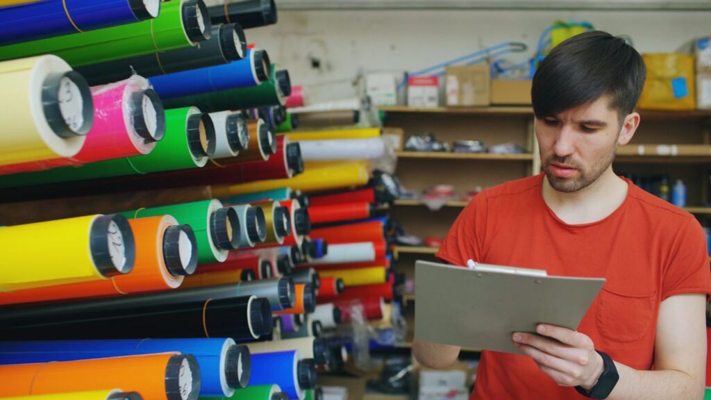 Man in a shop checking inventory of colorful paper rolls with a clipboard.